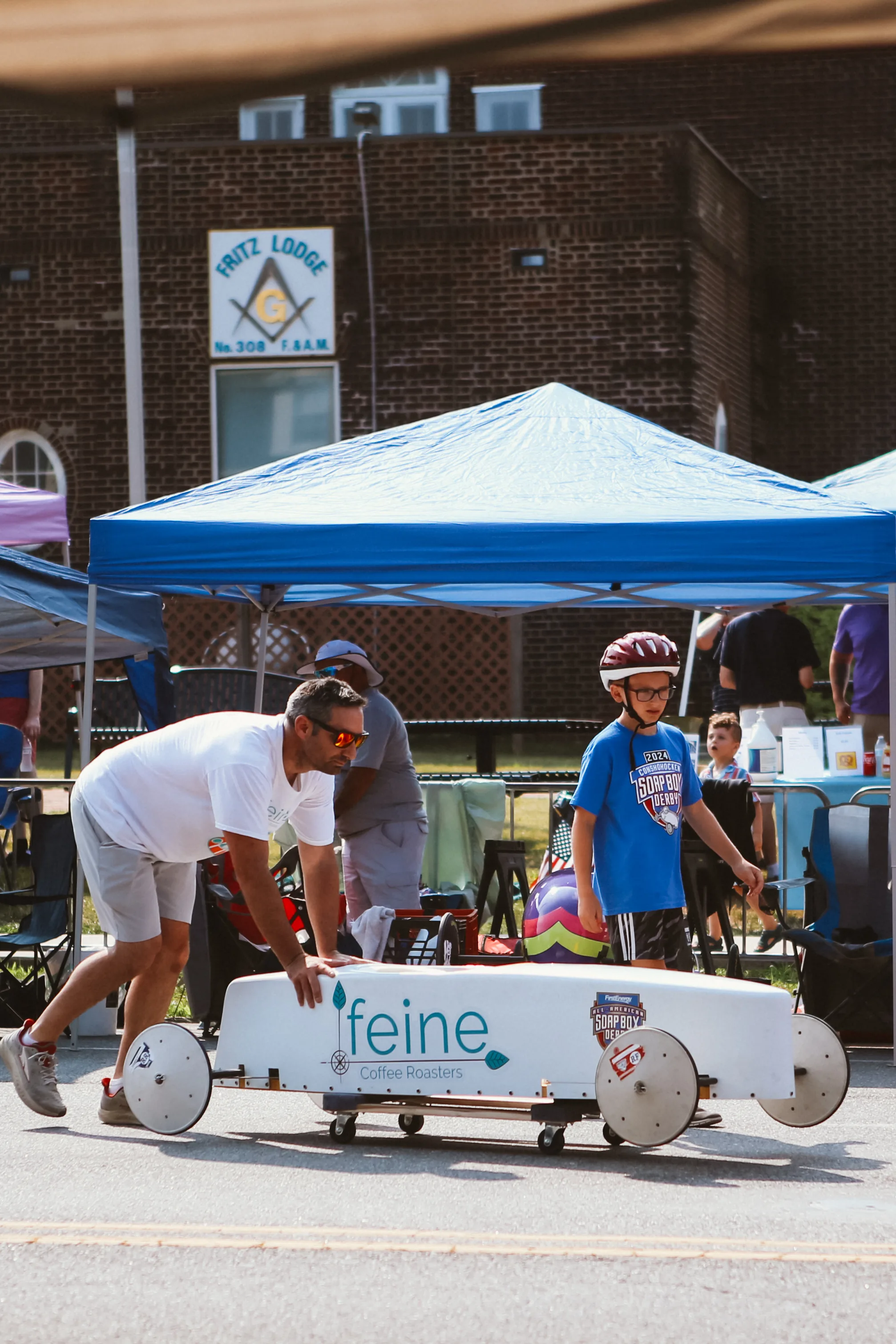 Soapbox derby scene in Conshohocken with a child in a race helmet and a derby car being guided down the street.